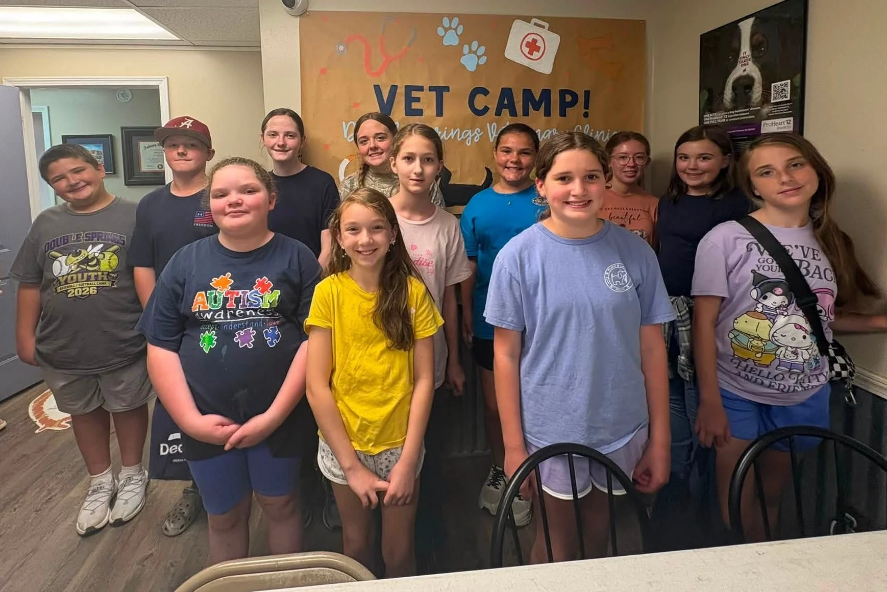 Vet Camp participants standing in front of a handmade sign that says, "Vet Camp! Double Springs Vet Clinic" with two cats and two dogs painted along the bottom underneath the text.